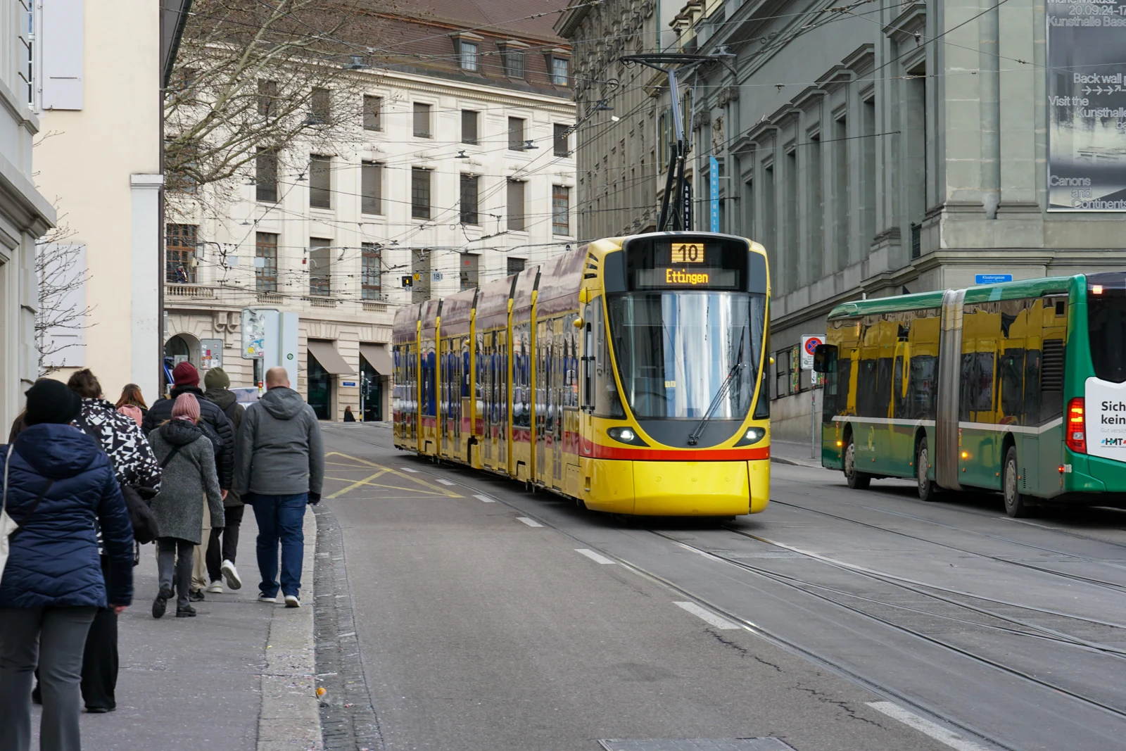 Şehir caddesinde ilerleyen sarı tramvay, yanındaki yeşil otobüs ve kaldırımdaki yayalar