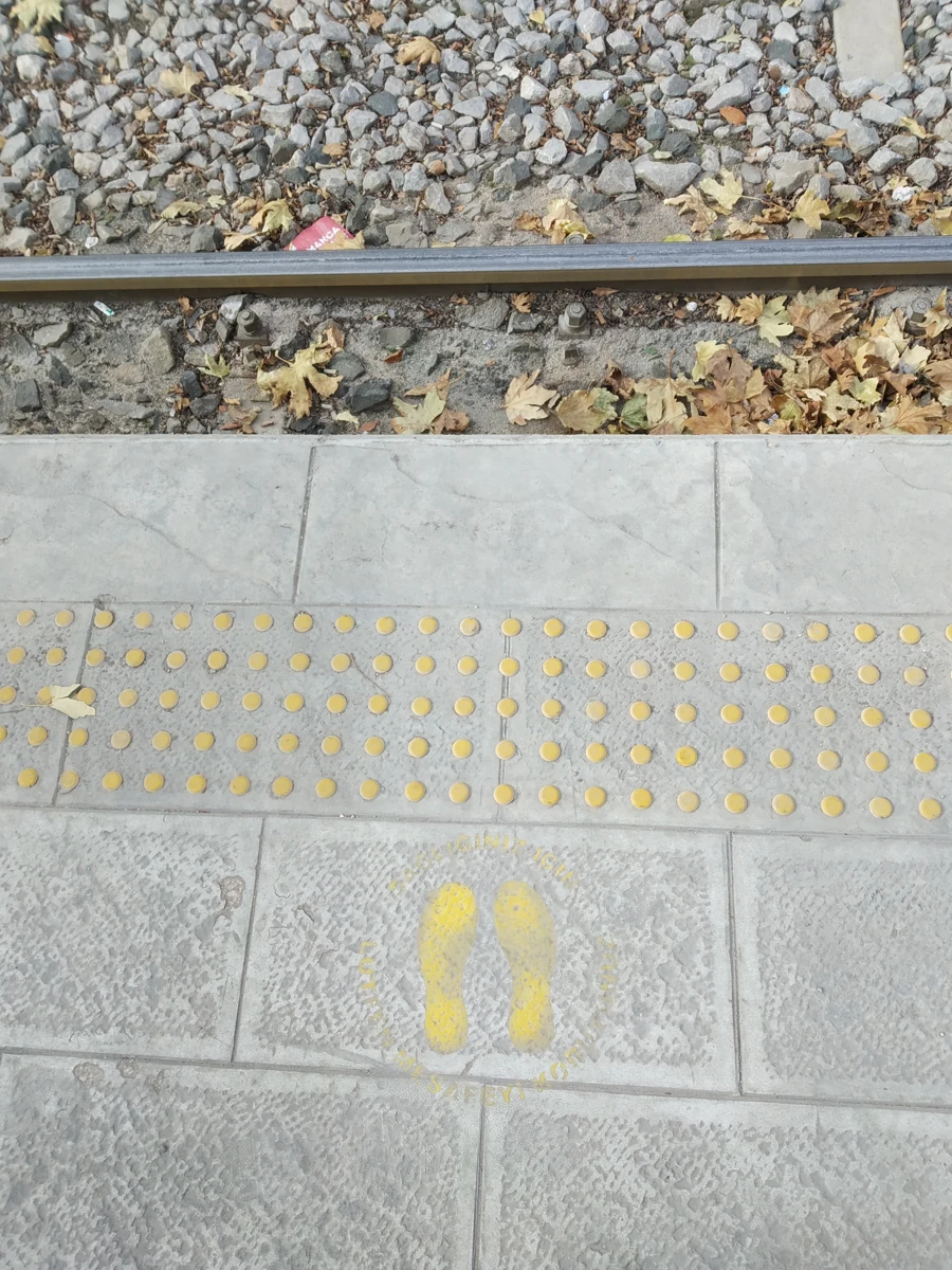 Tramvay rayı yanında sarı hissedilebilir yüzey ve ayak izi işareti bulunan kaldırım